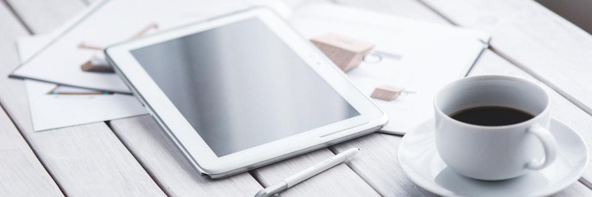 Een witte, houten tafel waar een wiite tablet en een witte kop koffie op liggen