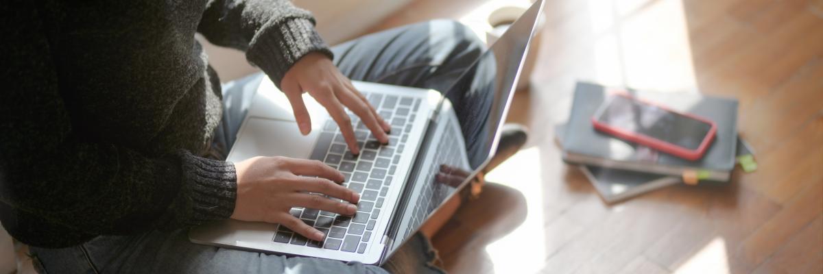 Vrouw die in kleermakerszit op haar laptop bezig is, haar telefoon ligt naast haar.