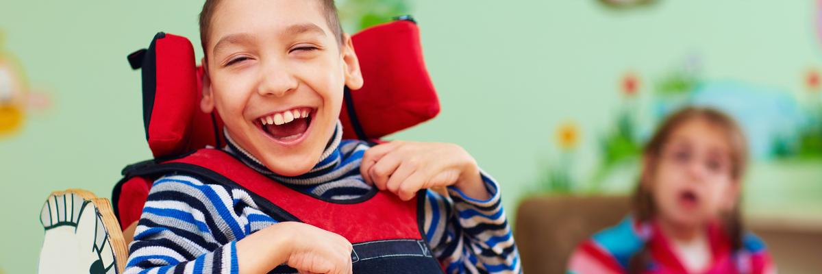 Een jongen met een lichamelijke beperking zit in zijn rolstoel en glimlacht breed naar de camera.