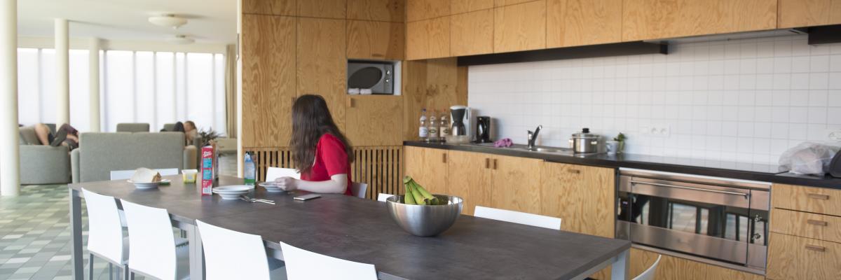 Een moderne keuken met veel wit en hout. Aan de keukentafel zit een jonge vrouw met een rode trui.