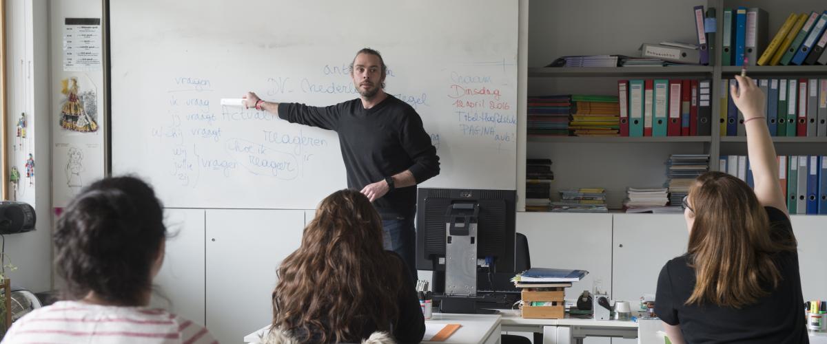 Een klaslokaal waar de leerkracht aan een whiteboard staat terwijl een van de meisjes haar hand opsteekt.