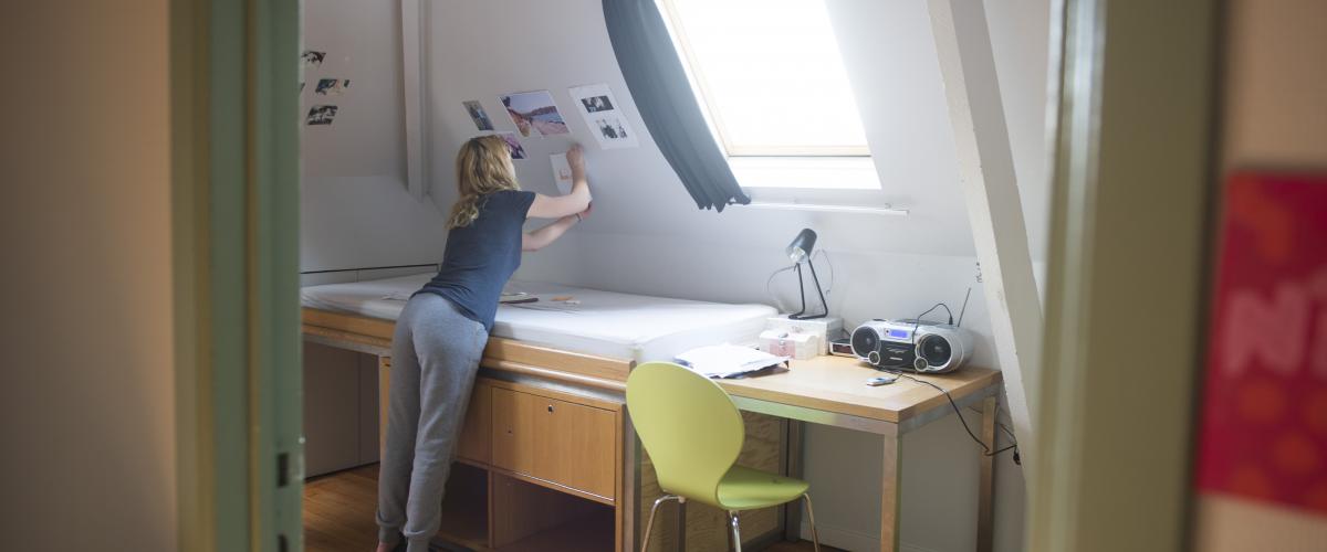 We zien door een deuropening een meisje een poster ophangen in haar kamer. Ze buigt over haar bureau om aan de schuine kant van de muur te komen.