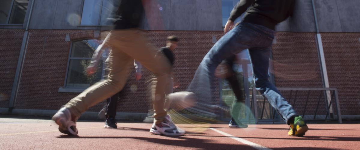 Verschillende jongeren die buiten voetballen op het basketbalterrein. De foto is zo getrokken dat je de bewegingen kan zien.