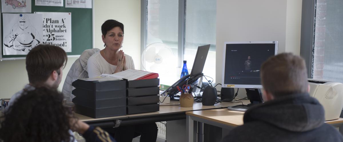 Een docent is les aan het geven in haar lokaal. De jongeren zitten met hun rug naar de camera. De docent is een uitleg aan het geven waarbij ze haar handen tegen elkaar heeft, alsof ze bidt.