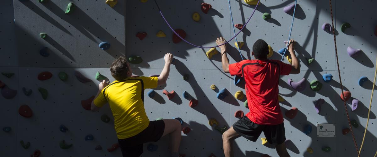 Twee jongeren, een met een geel t-shirt, de andere met een rood, klimmen op de klimmuur. De zon schijnt naar binnen waardoor de jongens nog meer opvallen.