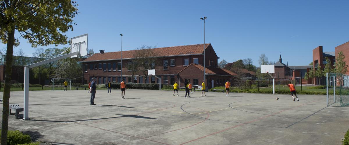Een groot basketbalveld buiten waar enkele jongeren aan het voetballen zijn. Op de achtergrond zie je een bijgebouw.