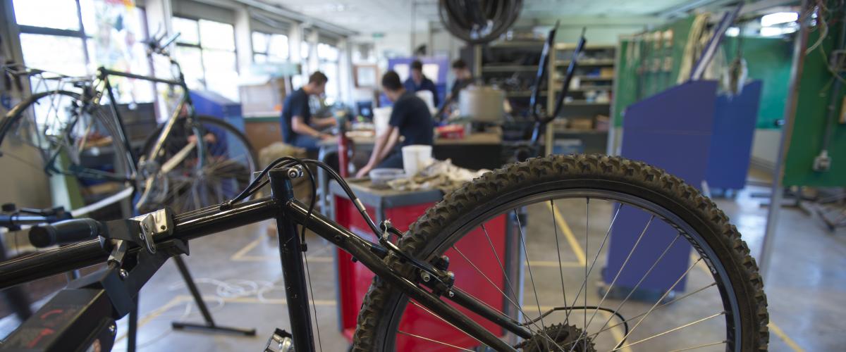 Een klaslokaal waar fietsen worden gerepareerd. Op de voorgrond zie je een fiets ondersteboven hangen. Achteraan zie je de jongeren die van de docent les krijgen.