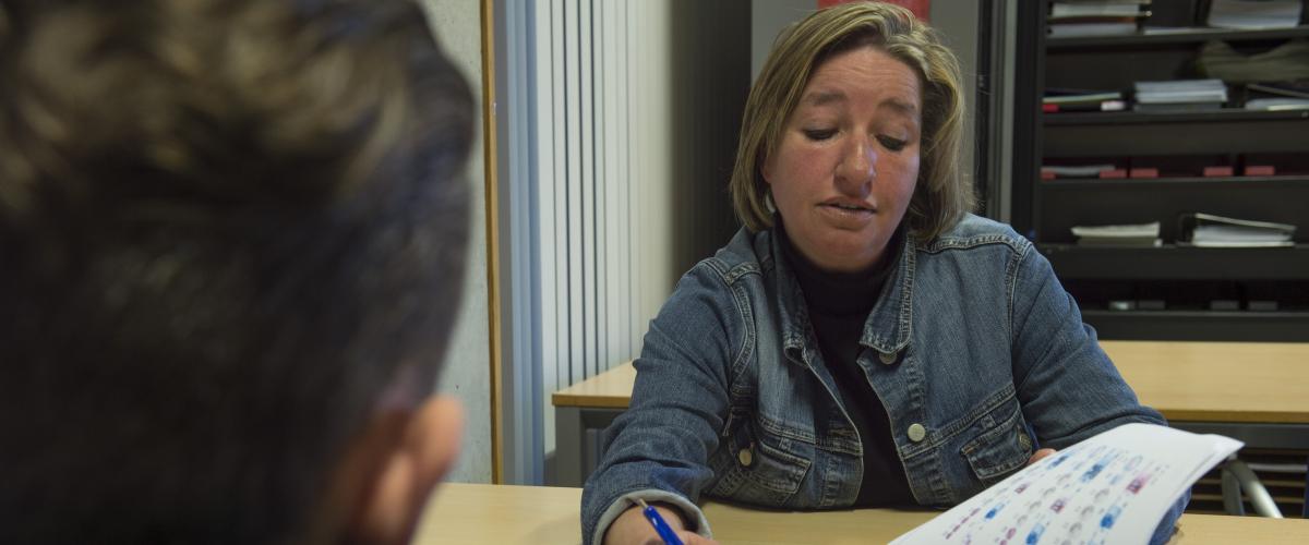 Een docente die in een boek bladert en iets aanwijst voor de jongere die voor haar aan tafel zit. De jongere zit met zijn rug naar de camera.