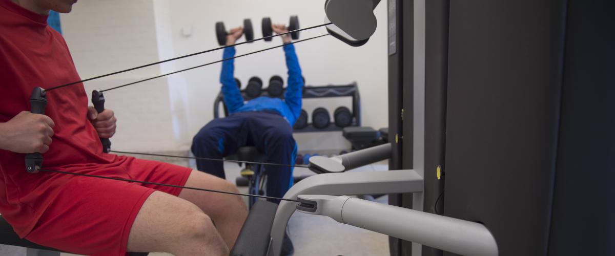 Een jongere in een rood sport tenue dat op een fitness machine oefeningen doet. Hij trekt aan kabels met gewichten.