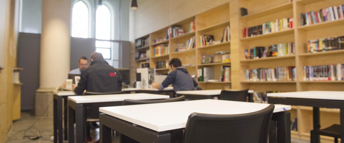 Een klaslokaal met ingebouwde boekenkasten. We zien twee jongeren aan een tafel die les krijgen van een docent.