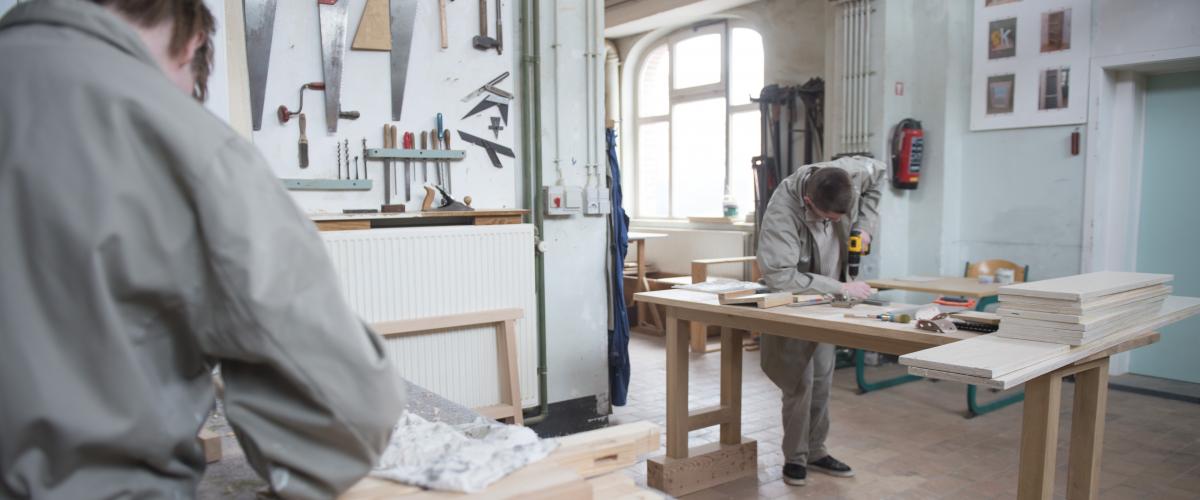 Een lokaal waar houtbewerking wordt gedaan. Twee jongeren zijn bezig met het zagen van een blok hout.