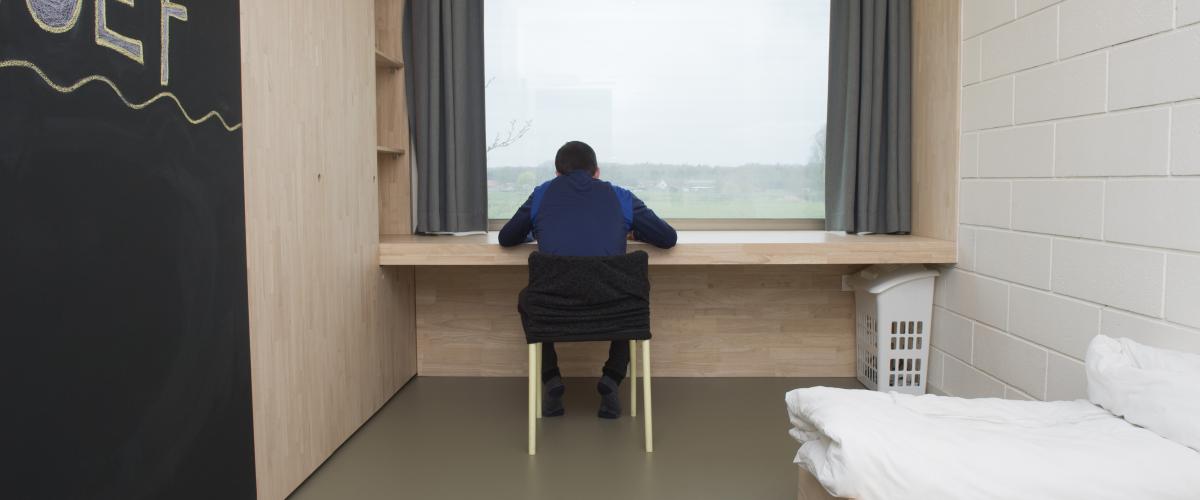 Een jongere die aan zijn bureau zit in zijn slaapkamer. Hij zit met zijn gezicht naar het raam, zijn bed aan zijn rechterzijde en een kalkbord aan zijn linkerzijde. op het bord staat 'Oef' geschreven.