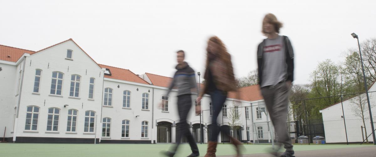 Drie jongeren, twee jongens en een meisje wandelen over de binnenplaats. Het hoofdgebouw en het sportterein op de achtergrond.