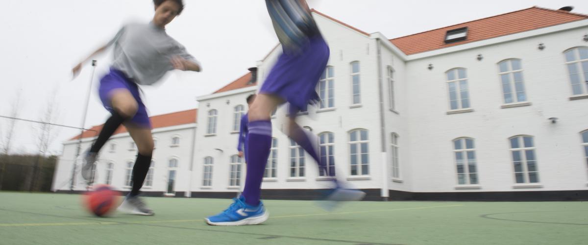 Twee jongeren in sporttenue die buiten voetballen. Het witte bijgebouw staat te pronken op de achtergrond.