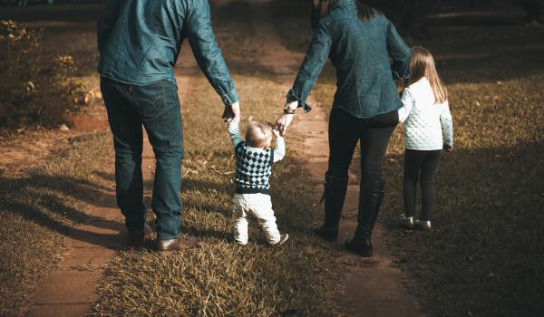 Familie van 4, houden hand vast. Jongste telg wandelt hobbelig, terwijl ouders hand hem ondersteunen