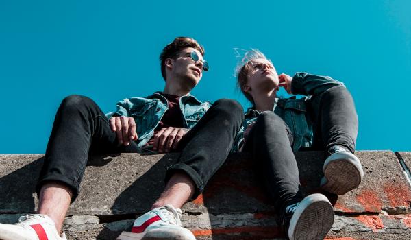 Een jonge man en vrouw, zittend op een muurtje, de foto is in kikkerperspectief getrokken zodat je de blauwe lucht ziet.