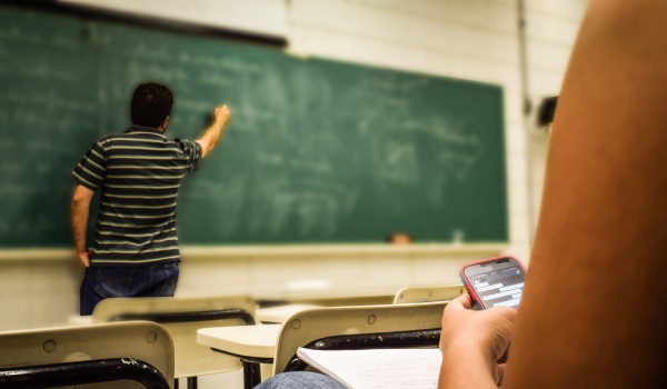 Een leerkracht schrijft op een schoolbord, we zien een studente met een gsm kijken.