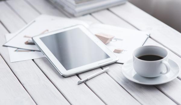 Een witte, houten tafel waar een wiite tablet en een witte kop koffie op liggen