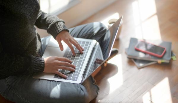Vrouw die in kleermakerszit op haar laptop bezig is, haar telefoon ligt naast haar.