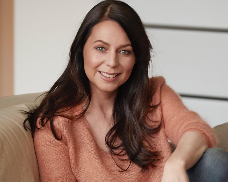 Kristel Verbeke, in een zetel met een lichte oranje trui en een gescheurde jeansbroek