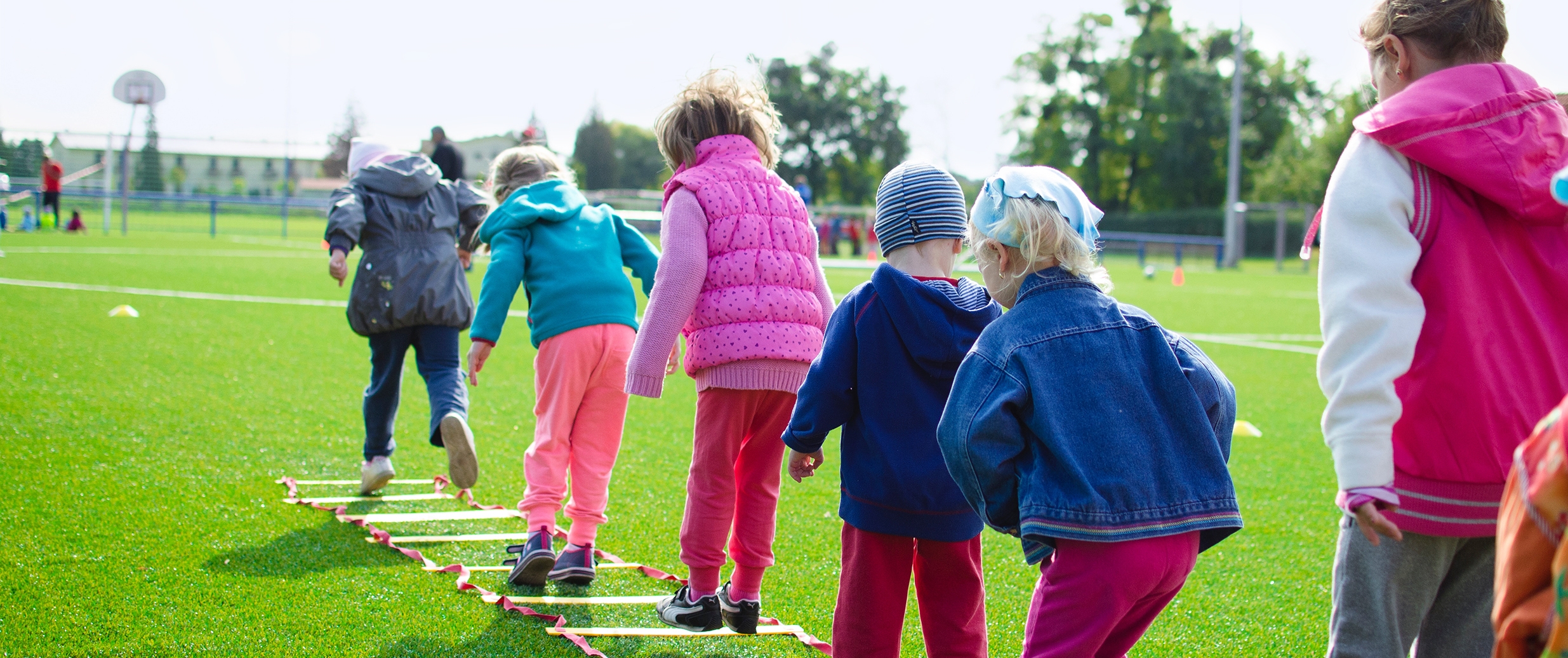 Een rij met kinderen die een springspel doen. Een ladder ligt in het gras en ze springen van vak naar vak.