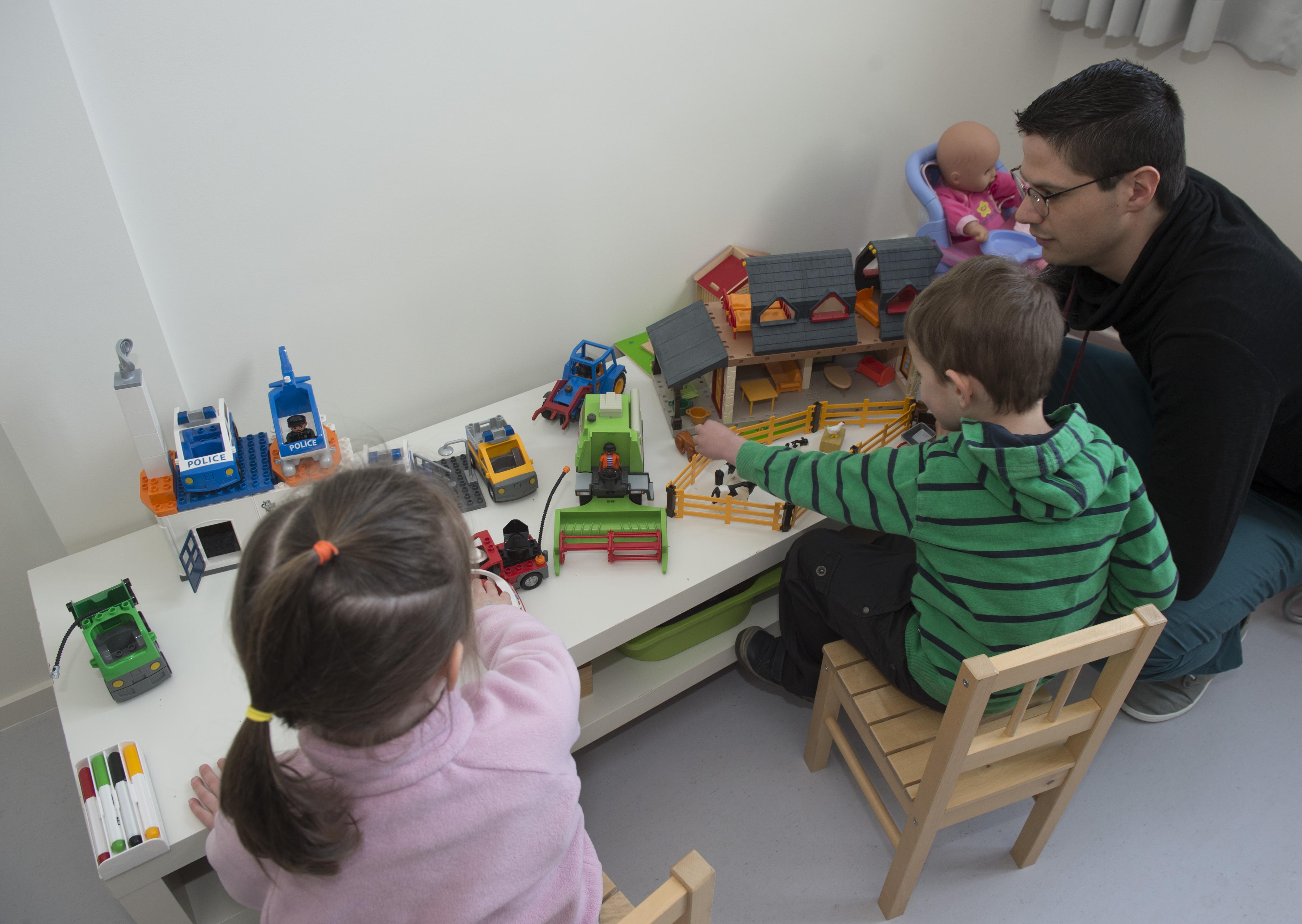 Een man die een jongen en een meisje observeert terwijl ze met speelgoed op een tafel spelen.